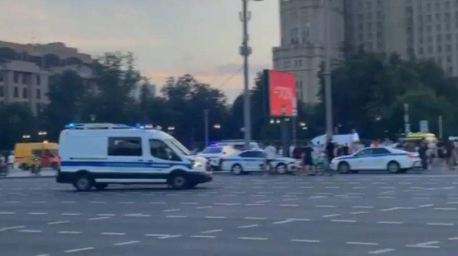 Policijska vozila sa uključenim rotacionim svjetlima na ulici, saobraćaj se odvija, prolaznici na trotoaru.
