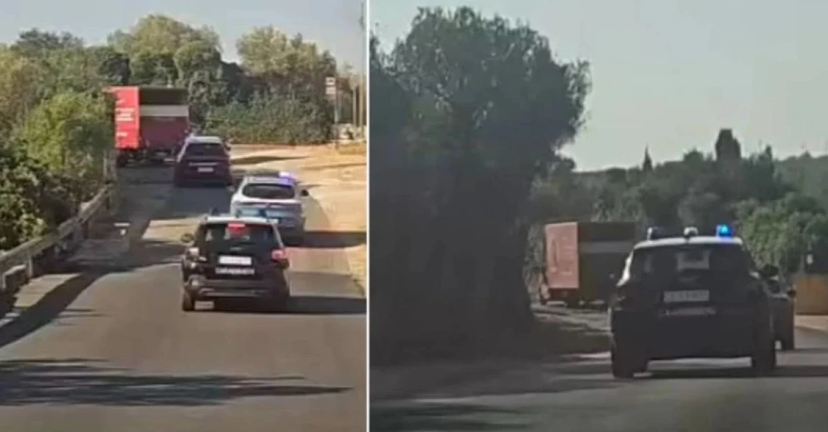 Crni policijski automobil s upaljenim plavim rotirkama prati kamion i druga vozila na putu.