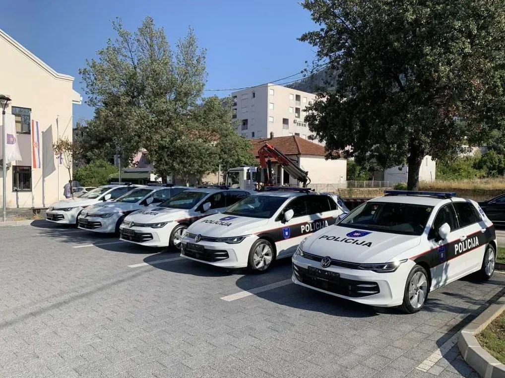 Nekoliko novih bijelih policijskih automobila marke Volkswagen Golf 8 parkiranih u nizu ispred zgrade policijske stanice, sa zastavama Hrvatske i zastavom Evropske unije na jarbolima.