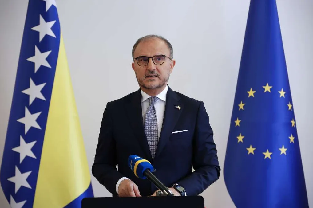 Muškarac u tamnom odijelu, bijeloj košulji i sa kravatom stoji ispred zastave Bosne i Hercegovine i zastave Evropske unije. Ispred njega se nalazi mikrofon.