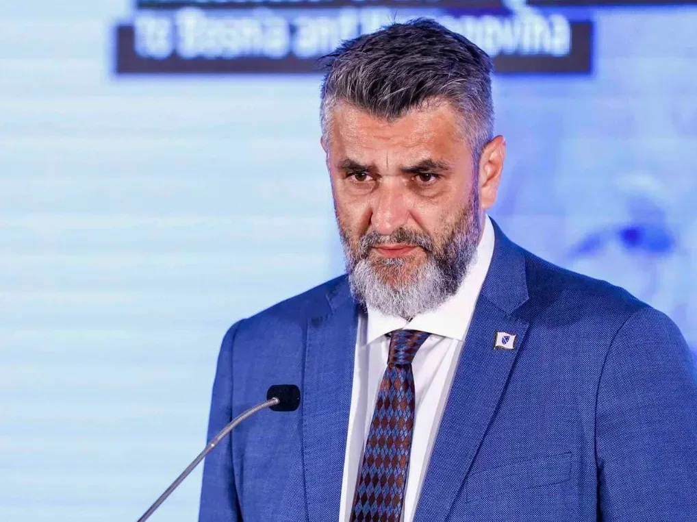 Političar sa bradom i plavim odijelom govori za govornicom.