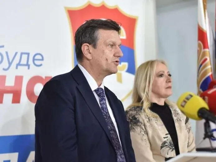 Muškarac u tamnom odijelu i sa plavom kravatom govori u mikrofon, dok pored njega stoji žena sa plavom kosom i jaknom bež boje sa crnim uzorkom. U pozadini se vide zastave i grb.