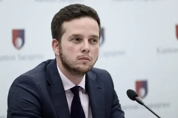 Portret mladog muškarca u tamnom odijelu i bijeloj košulji s tamnom kravatom, kako sjedi ispred pozadine s grbom Kantona Sarajevo.