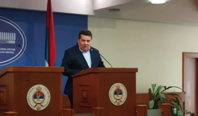 Čovjek u tamnom odijelu stoji za drvenim podijumom i govori.