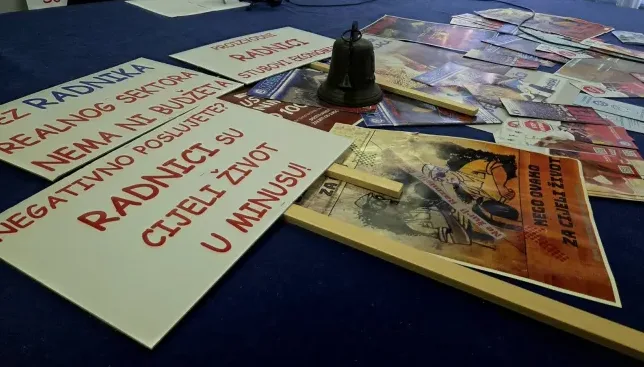 Fokus na bijele transparente sa crvenim natpisima koji izražavaju nezadovoljstvo radnika, a na jednom od njih stoji "Bez radnika realnog sektora, nema ni budžeta. Negativno poslujete, radnici su cijeli život u minusu!". U pozadini se vide drugi transparenti i bronzani zvonik.