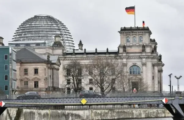 Reichstag zgrada sa kupolom i njemačkom zastavom na vrhu, sa mostom i saobraćajem u prednjem planu.