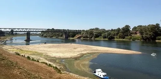 Prizor rijeke sa pješčanim sprudom u prvom planu, metalnim mostom u daljini i zelenilom na obalama.