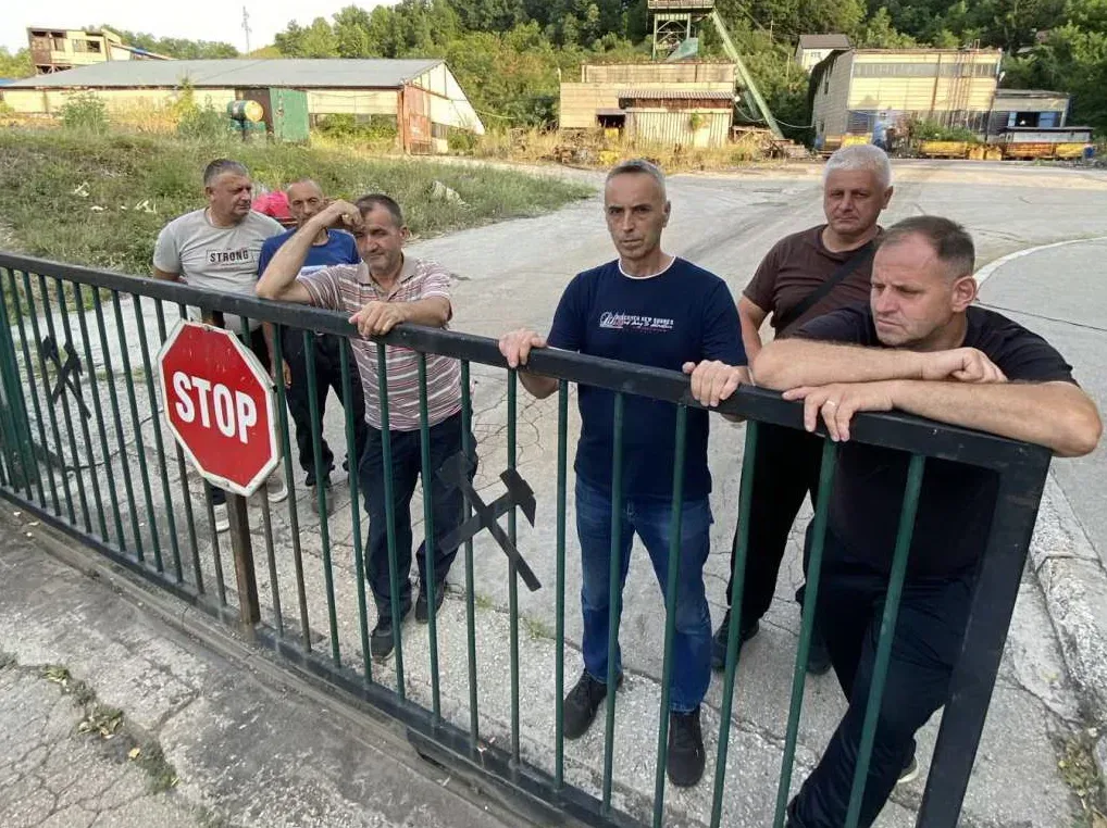 Grupa rudara stoji iza metalne ograde sa znakom 'STOP' ispred rudarskih objekata.