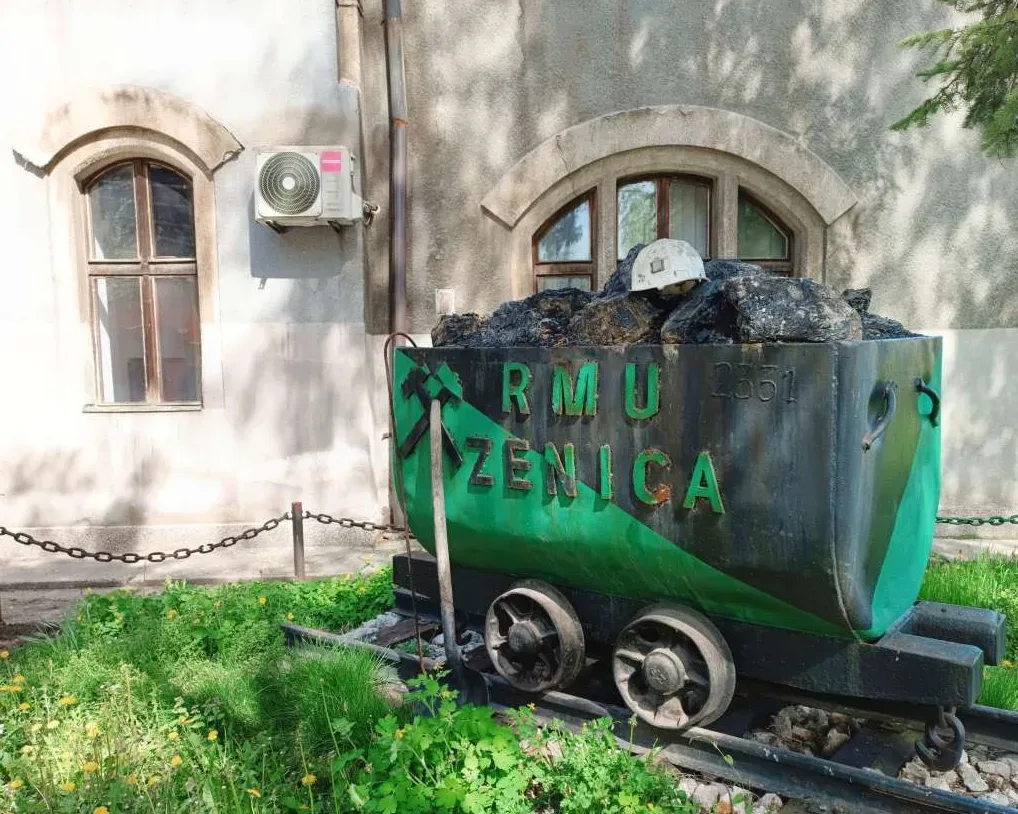 Rudarski vagon pun uglja sa kacigom na vrhu, ispred zgrade sa prozorima