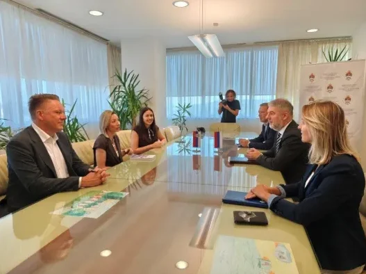Grupa ljudi sjedi za velikim sjajnim stolom u modernoj kancelariji, razgovarajući. Jedna osoba stoji iza njih i snima događaj kamerom.