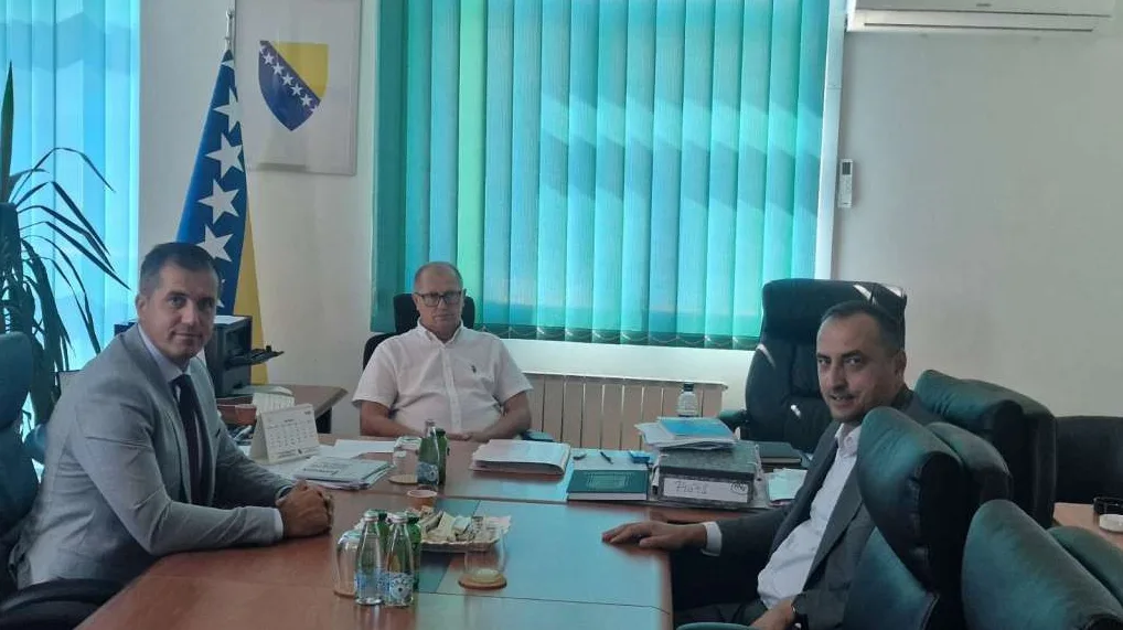 Tri muškarca sjede za stolom u kancelariji, jedan s lijeve strane gleda u kameru, a druga dvojica desno. U pozadini je zastava Bosne i Hercegovine i plave žaluzine.