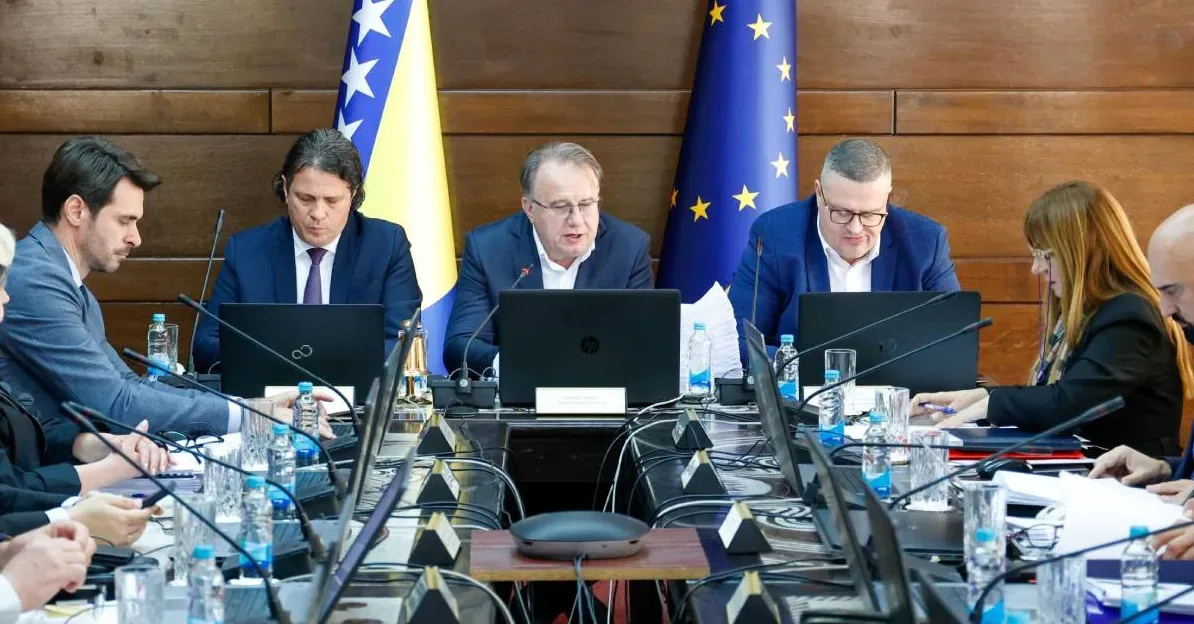 Zvaničnici sjede za stolom tokom sastanka, fokusirajući se na svoje laptope, sa zastavama Bosne i Hercegovine i Evropske unije u pozadini.