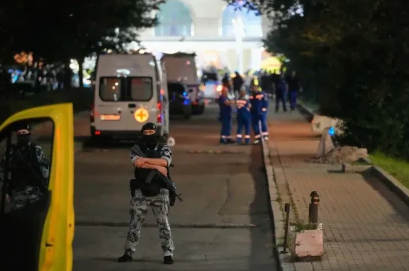 Dva naoružana vojnika u maskirnim uniformama i fantomkama stoje ispred žutog vozila noću. U pozadini se vide ambulantna vozila i osobe u plavim uniformama, vjerovatno medicinsko osoblje ili policija, te zgrada u daljini.