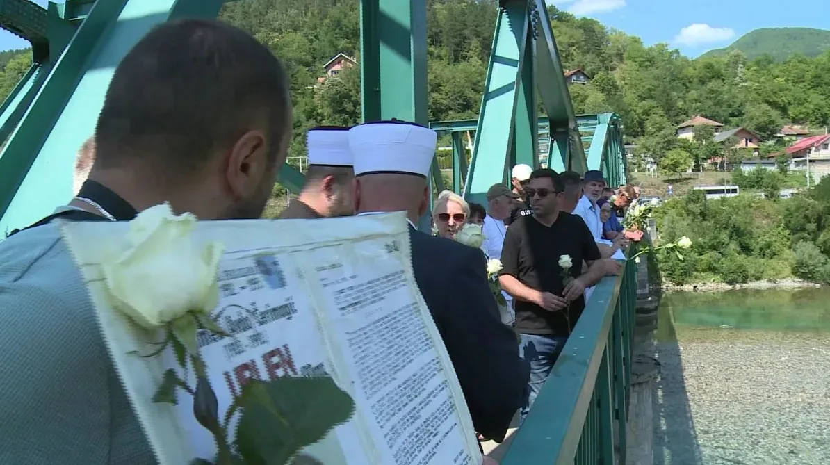 Ljudi na zelenom mostu drže bijele ruže i dokument, prisjećajući se događaja.