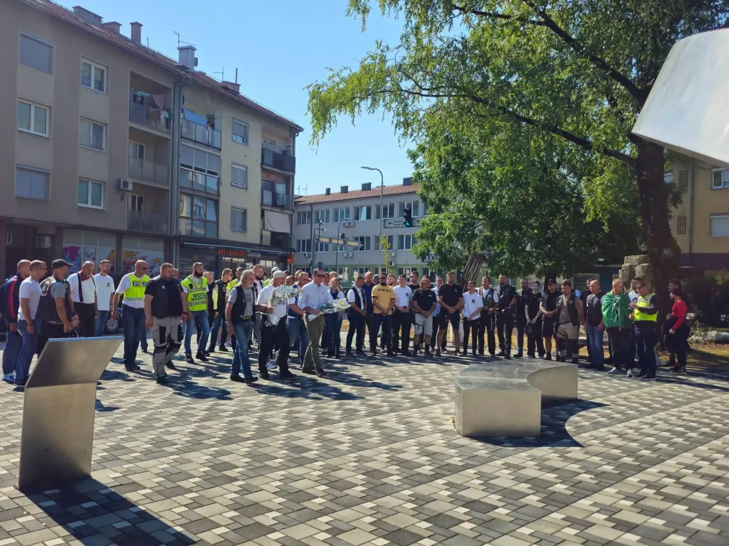 Grupa ljudi, među kojima ima i policajaca u uniformi i civilnim odijelima, kao i motociklista, okupljena je na popločanom trgu na sunčan dan.