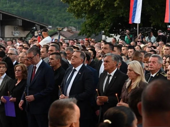 Veliki broj ljudi na skupu drži upaljene svijeće i gleda u jednom pravcu, u pozadini se vide nacionalne zastave i drveće.