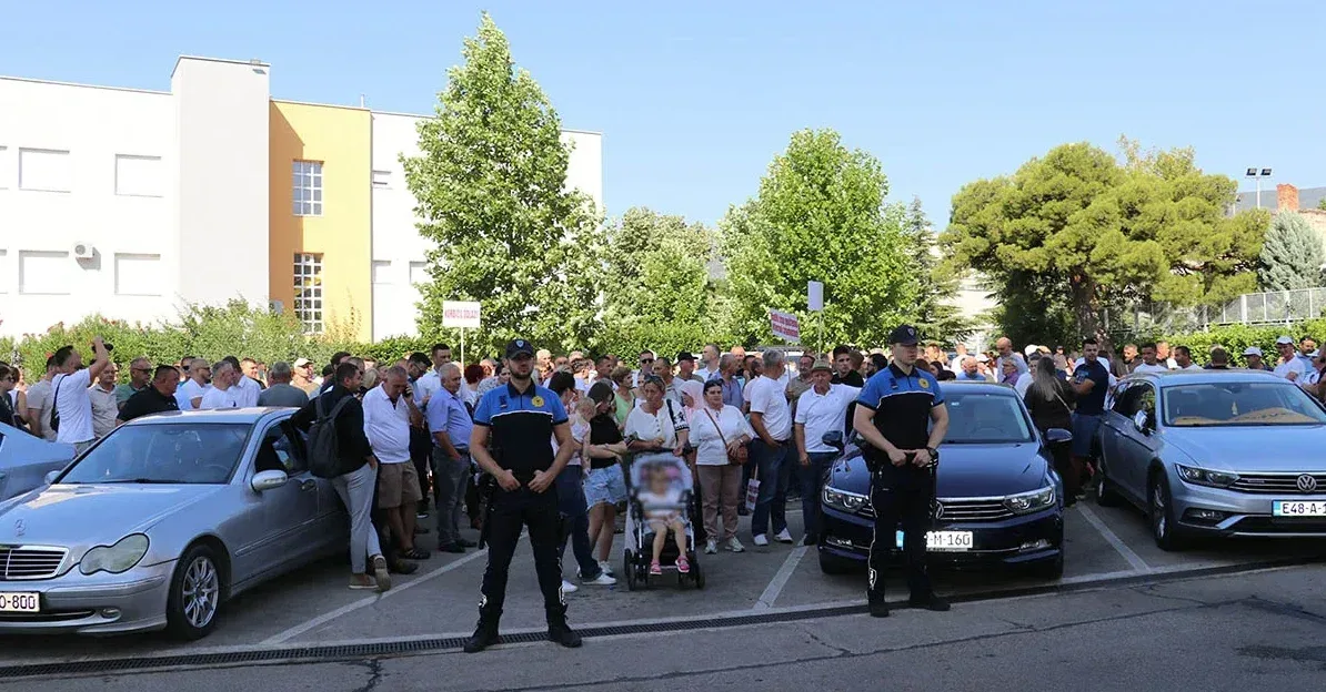 Grupa ljudi stoji ispred parkiranih automobila dok policajci obezbjeđuju skup.