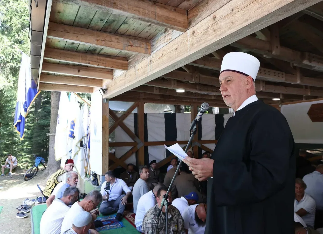 Imam u bijelom turbetu i crnom odijelu drži papir i govori u mikrofon, dok mu u pozadini sjede vjernici na prostirkama i zastave Bosne i Hercegovine.