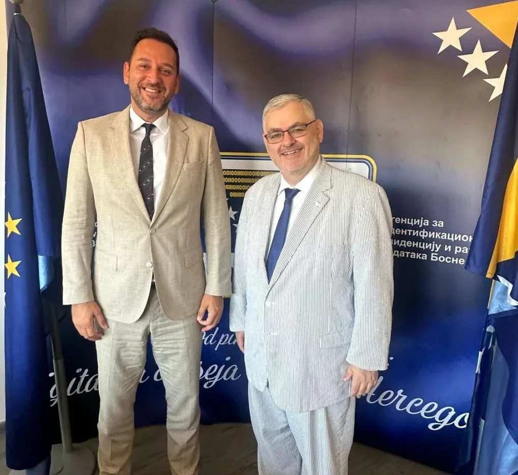 Dva muškarca u odijelima stoje ispred zastava i natpisa.