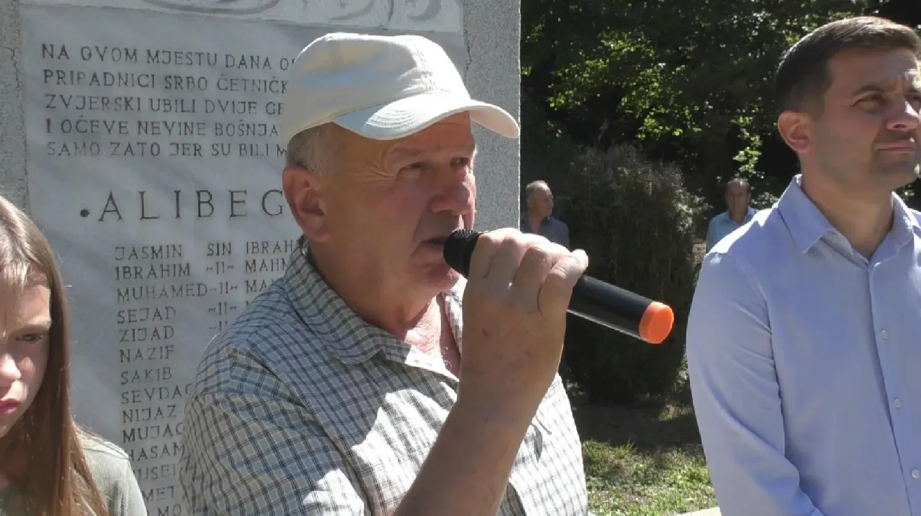 Čovjek drži mikrofon ispred spomen-ploče s imenima i natpisom o ubijenim Bošnjacima.