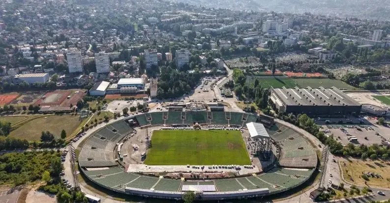 Sportski stadion sa zelenim terenom i tribinama okružen gradskom okolinom.