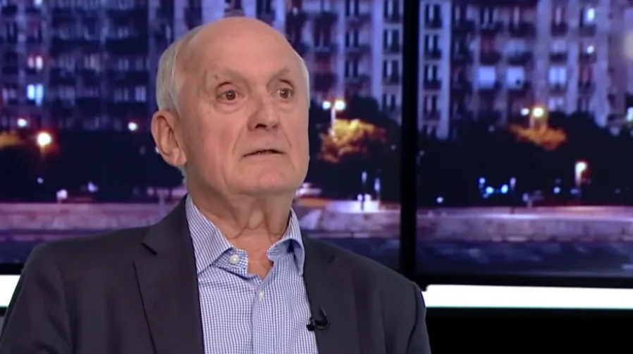 Stariji gospodin sa kratkom sijedom kosom, odjeven u tamno odijelo i plavu kariranu košulju, sjedi u televizijskom studiju i govori.