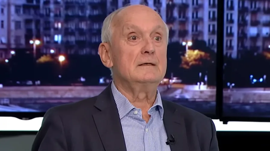 Portret starijeg muškarca sa sijedom kosom, obučenog u tamni sako i plavo-bijelu kariranu košulju, kako govori u studiju sa pozadinom koja prikazuje gradski pejzaž noću.