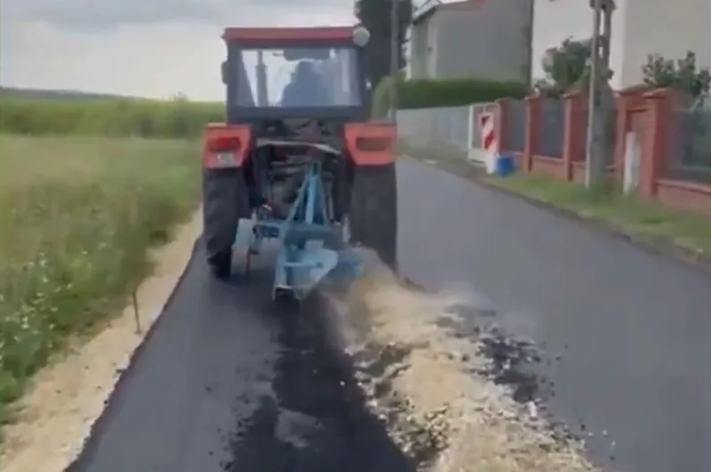 Crveni traktor sa prikačenom mašinom za asfaltiranje radi na novoasfaltiranom putu pored polja i stambenih objekata.