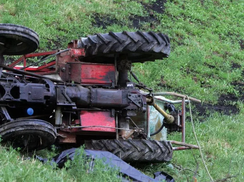 Prevrnuti crveni i crni traktor leži na zelenoj livadi, sa velikim gumama koje su iznad ostalih dijelova mašine.