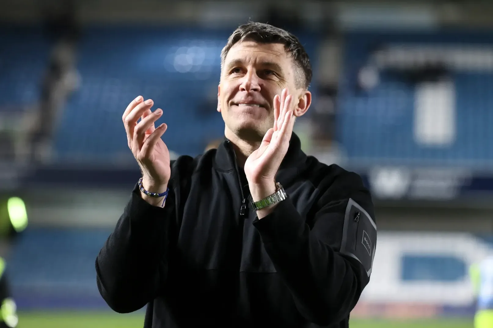 Fudbalski trener u crnom duksu aplaudira na stadionu.