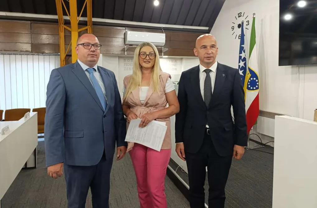 Dva muškarca u tamnim odijelima i žena u bež prsluku i roze pantalone stoje zajedno.