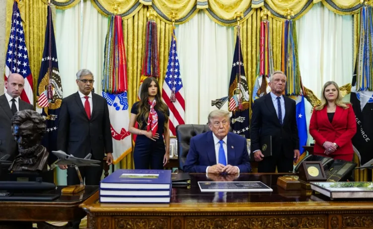 Donald Trump sjedi za stolom u Ovalnom kabinetu, okružen ljudima i zastavama, potpisuje dokumente.