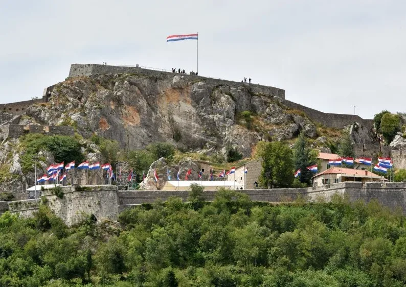 Pogled na staru kamenu tvrđavu na vrhu stjenovite planine, ukrašenu brojnim hrvatskim zastavama, dok se sa vrha tvrđave vijori velika hrvatska zastava.