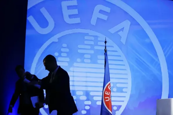 Siluete dvojice muškaraca u odijelima, jedan od njih prolazi pored zastave sa logom UEFA-e, dok se u pozadini vidi veliki ekran sa istim logom.