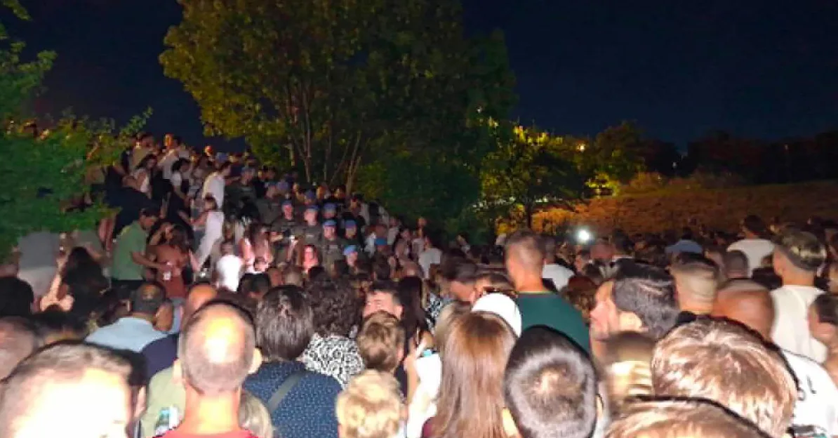Velika grupa ljudi okupljena na otvorenom prostoru tokom noći, dok se u pozadini vide stabla i osvijetljeni teren.