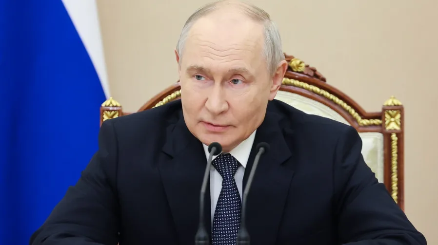 Vladimir Putin sjedi za stolom ispred dva mikrofona, s ruskom zastavom u pozadini.