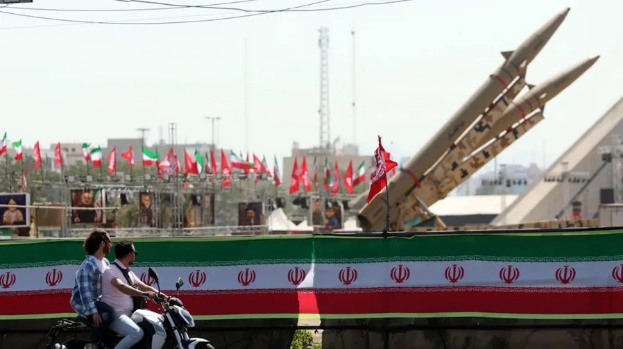Dvojica muškaraca na motociklu prolaze pored vojne parade na kojoj su izložene rakete, iranske zastave i portreti.