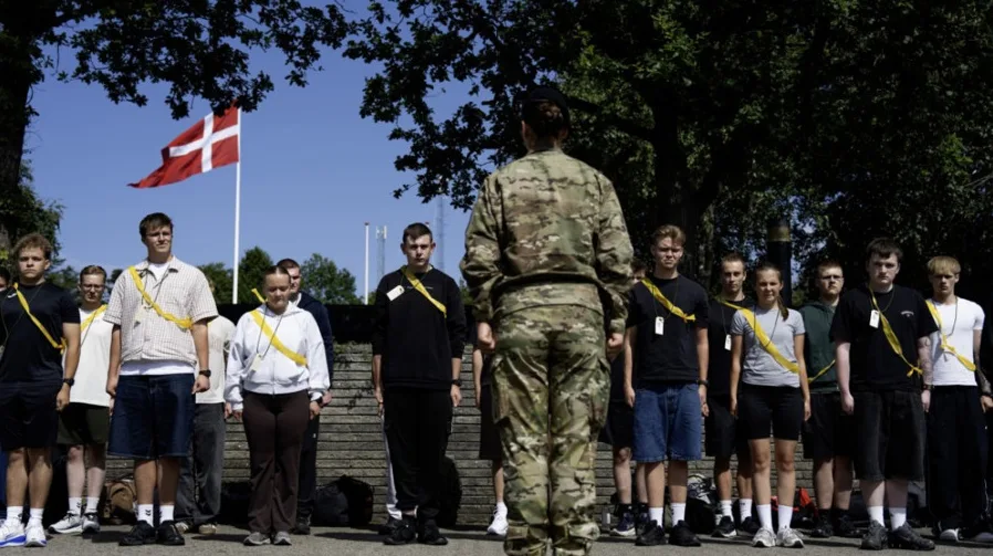 Grupa mladih ljudi u vojnim uniformama stoji u redu ispred danskog barjaka.