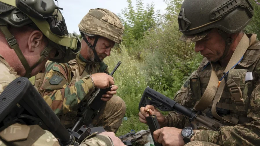 Vojnici u vojnim uniformama i kacigama pažljivo sastavljaju svoje automatske puške i municiju na otvorenom, okruženi vegetacijom.