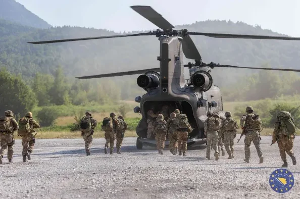 Vojnici u punoj opremi, s rancima na leđima, organizovano ulaze u otvoreni dio velikog vojnog helikoptera Chinook. Helikopter se nalazi na šljunčanom terenu, a u pozadini se vide zelene planine i drveće. Na desnoj strani fotografije nalazi se grb Evropske unije i Bosne i Hercegovine.