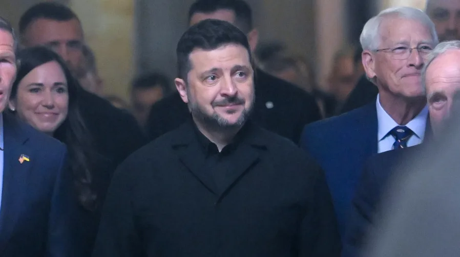 Volodimir Zelenski, ukrajinski predsjednik, u crnoj košulji i sakou, nasmijan, u pratnji američkog političara sa naočalama i plavim odijelom, te drugih osoba.