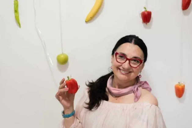 Žena sa crvenim naočarima i ružičastom maramom oko vrata drži crvenu papriku, okružena voćem i povrćem koje visi na koncima.