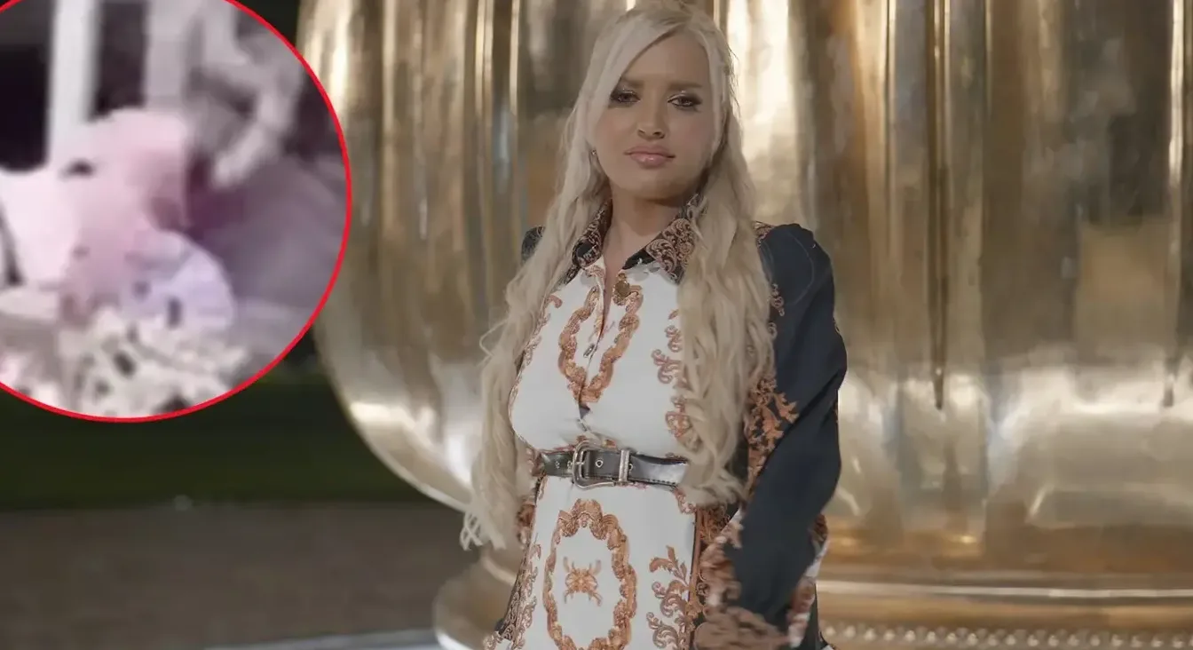 Žena sa plavom kosom u bijeloj haljini sa zlatnim ukrasima i crnim pojasom.