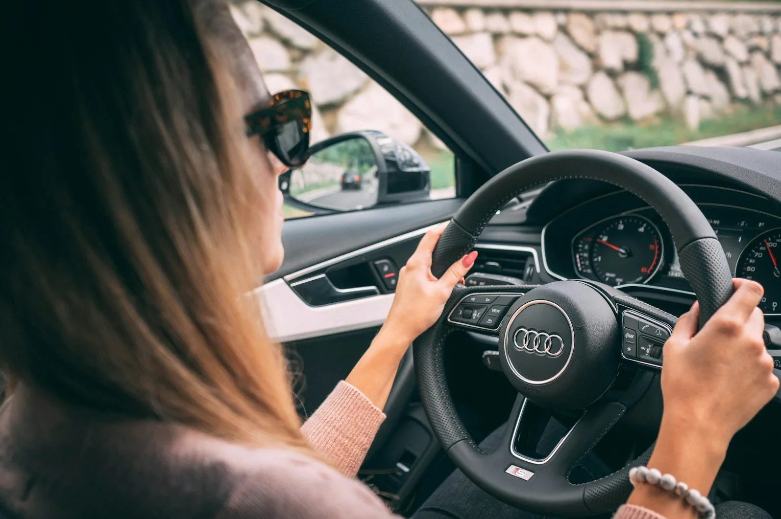 Žena sa dugom plavom kosom i naočarima za sunce vozi automobil marke Audi, držeći volan objema rukama.