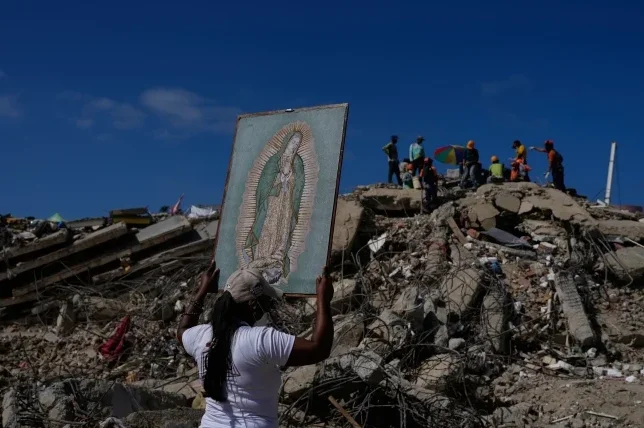 Žena drži sliku Gospe Guadalupske iznad glave, usred ruševina nakon zemljotresa. U pozadini se vide spasioci i razrušene zgrade pod vedrim nebom.